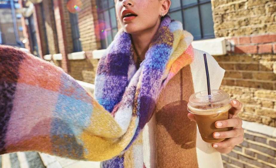 Woman outdoors on a city street holding an iced coffee; colorful plaid scarf, tan sweater, brick buildings and daylight enhance the scene.