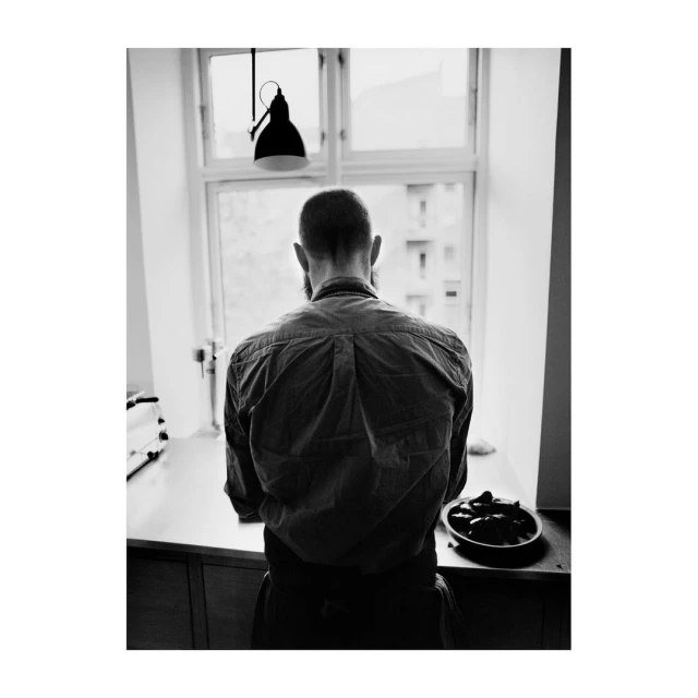 Black and white portrait from behind of an adult male with a beard standing in front of a window next to a countertop.