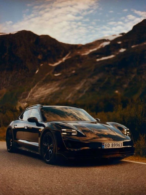 A black Porsche Taycan Cross Turismo is parked on an asphalt road with a backdrop of mountains and cloudy skies.