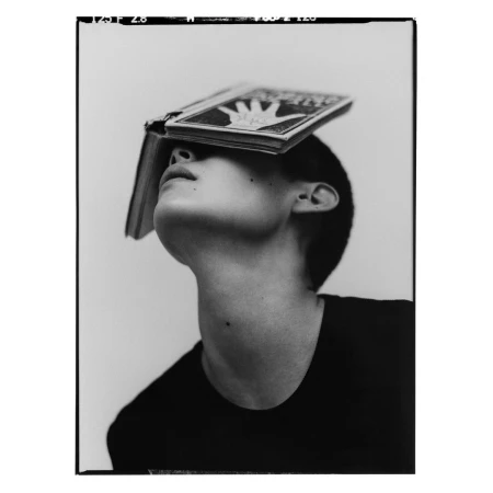 Monochrome portrait of a young adult with short hair wearing a dark shirt, balancing a book on their forehead while looking up.