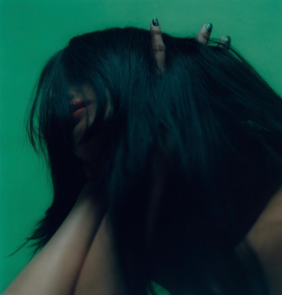 Portrait of a woman with long, dark hair against a green background, partially obscuring her face with her hands and hair.