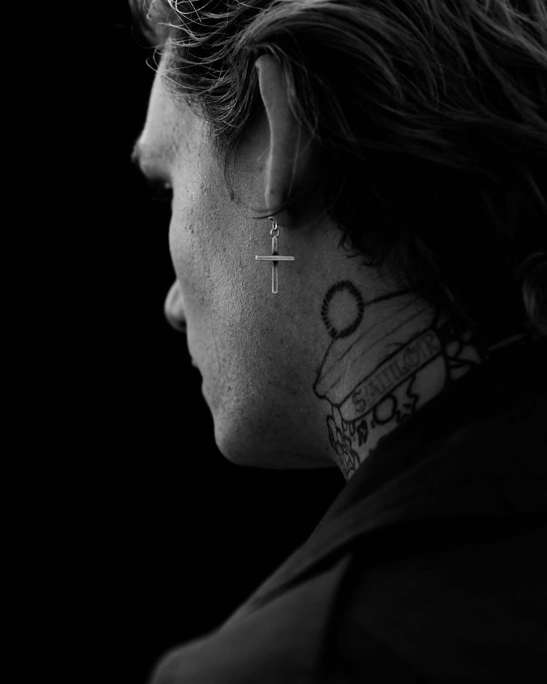 Monochrome portrait of adult male from the side, showcasing a cross earring and neck tattoo against a dark background.