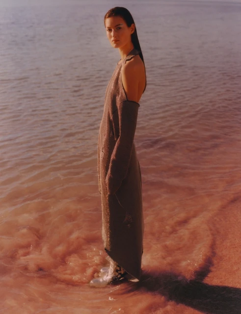 A full-length shot of a woman in a dress standing in shallow pink water, looking at the camera with a serene expression in a dreamy style.