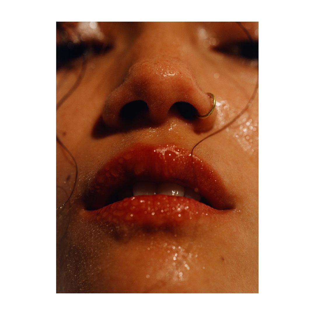 Close-up of a woman's face featuring shiny red lipstick, a nose ring, and wet skin, captured with a shallow depth of field.