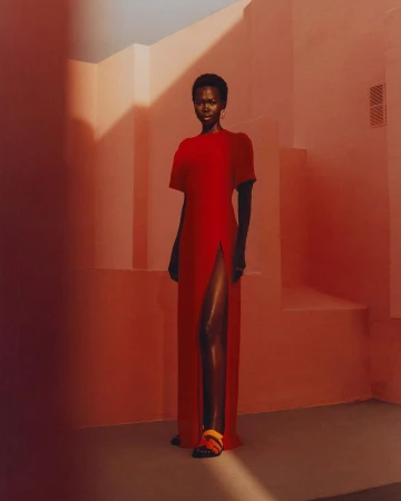 A black female model standing in a long red dress against a warm pink geometric architectural background photographed by Rory Payne.