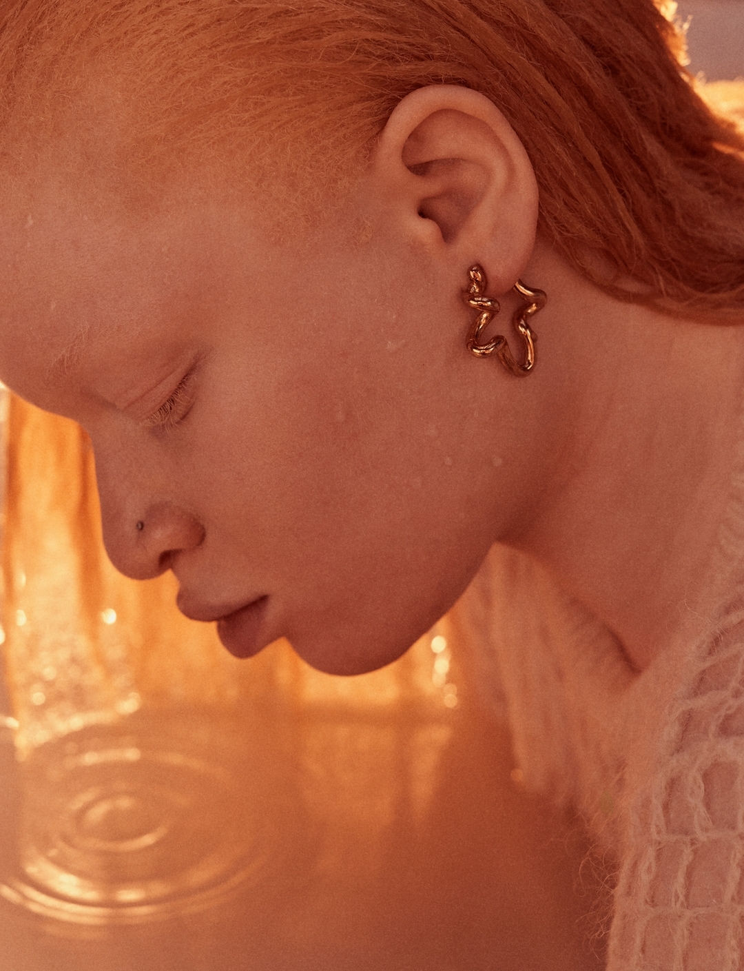 Close-up of an adult female with unique bronze earrings, fair skin, and short ginger hair, basking in soft, diffused light indoors.
