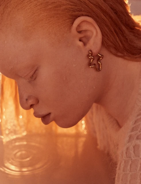 Close-up of an adult female with unique bronze earrings, fair skin, and short ginger hair, basking in soft, diffused light indoors.