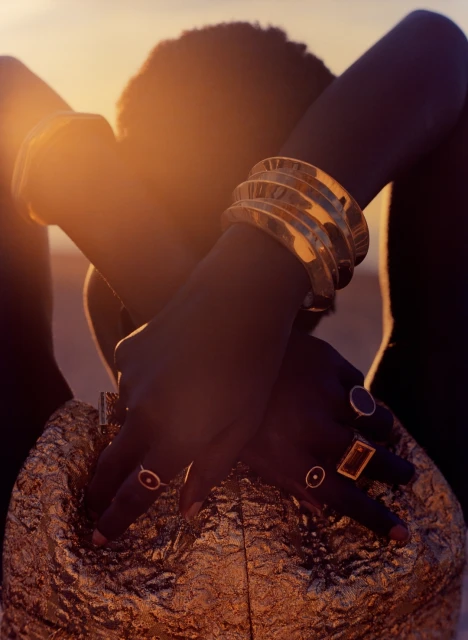 Close-up shot of a dark-skinned person with arms crossed, adorned with gold bracelets and rings, and wearing a golden, textured top.