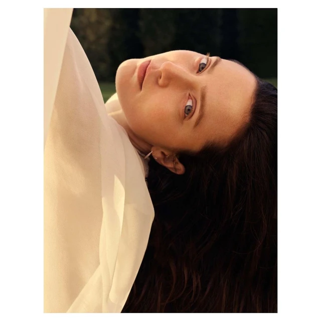 Adult woman gazes upwards with a slight smile; wearing a flowing white garment against a blurred outdoor background; natural light.