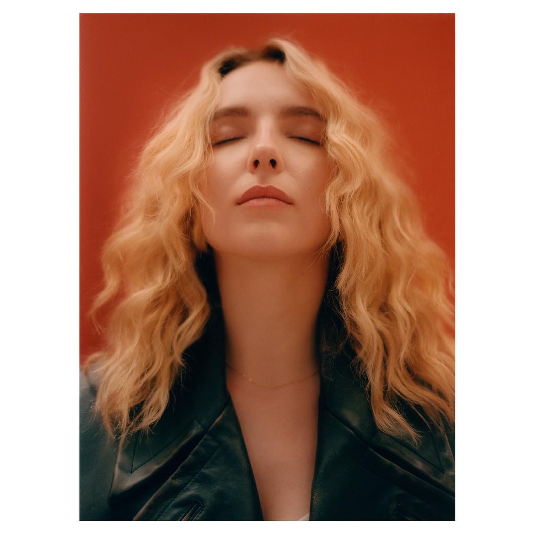 Portrait of a blonde woman with her eyes closed wearing a dark jacket against a solid red backdrop photographed by Rory Payne.