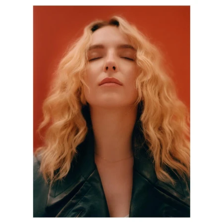 Portrait of a blonde woman with her eyes closed wearing a dark jacket against a solid red backdrop photographed by Rory Payne.