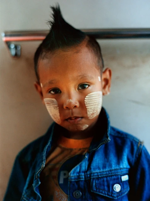 Close-up portrait of a young child with a mohawk hairstyle, painted facial markings, and a denim jacket indoors, eye-level angle.
