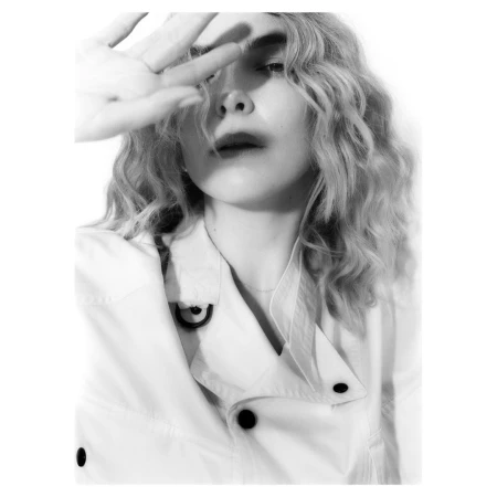Close-up portrait of a woman with wavy blonde hair holding her hand up near her face, wearing a white collared jacket.