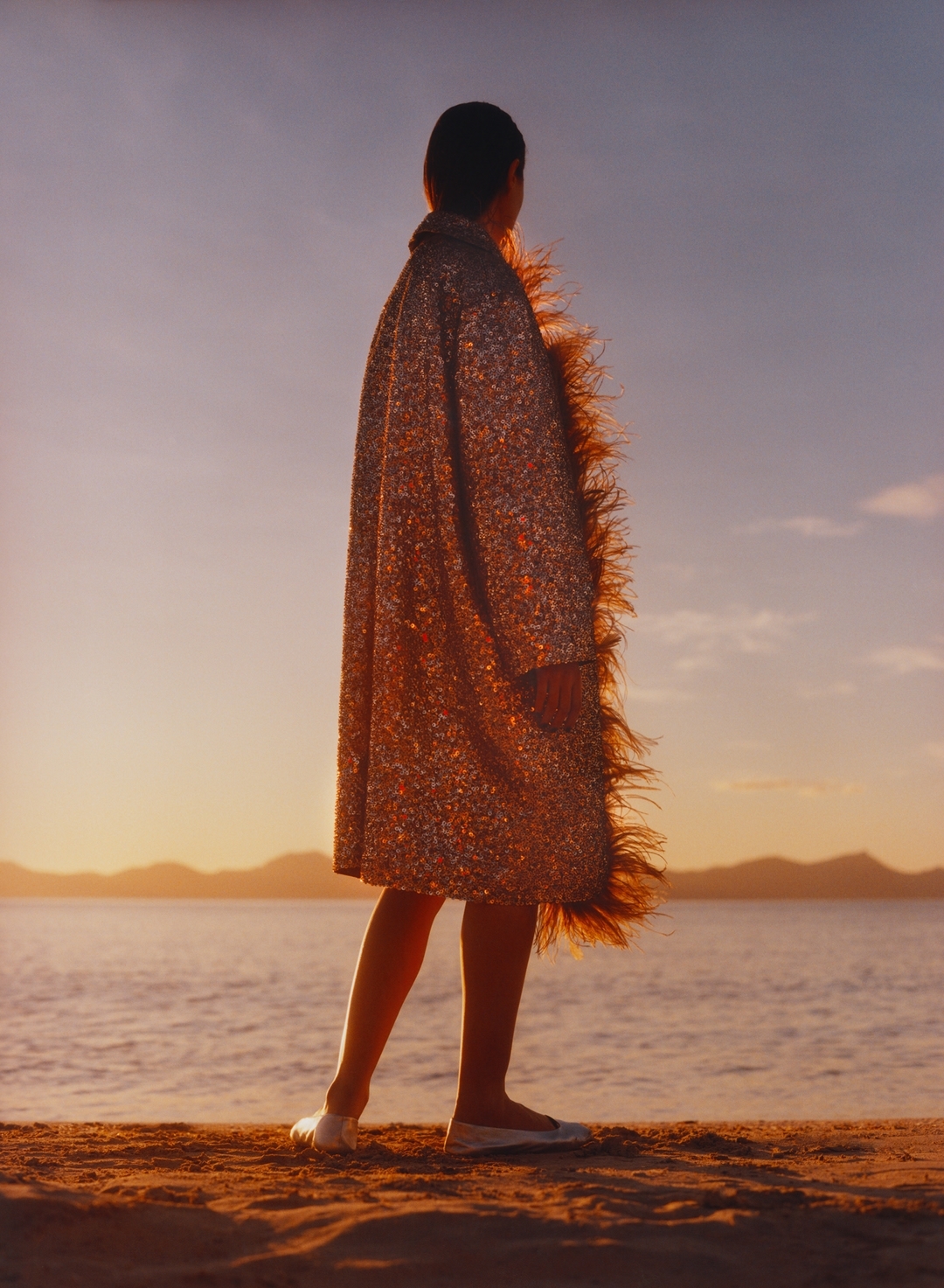 A woman stands on a sandy beach at sunset, wearing a sparkly coat with feather trim, facing away from the camera, toward the ocean.
