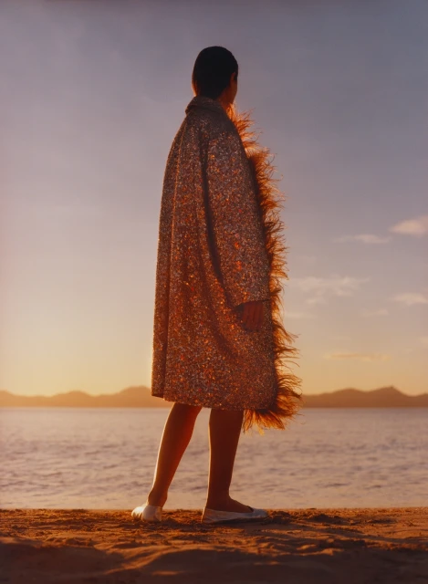 A woman stands on a sandy beach at sunset, wearing a sparkly coat with feather trim, facing away from the camera, toward the ocean.