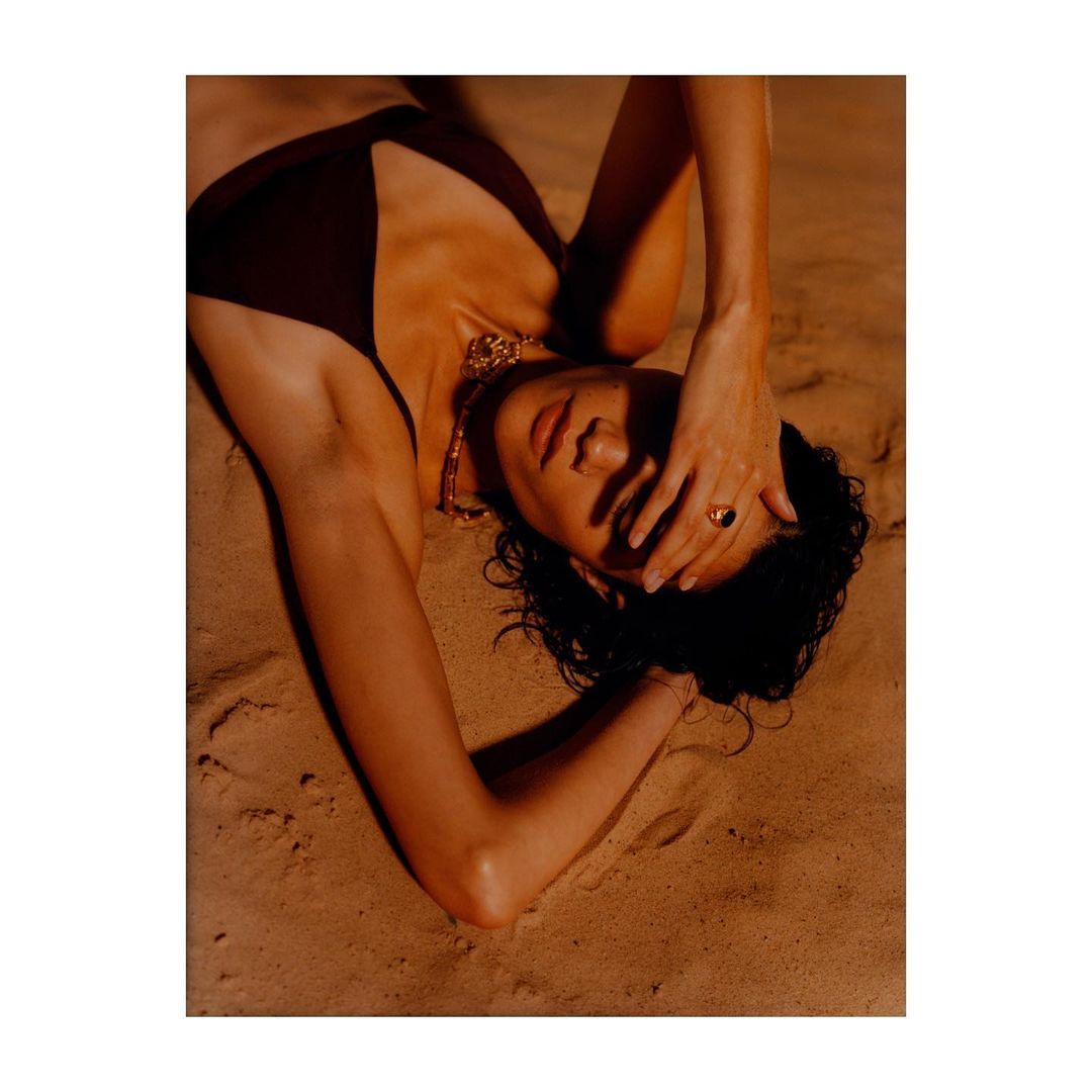 Model lying on sandy terrain wearing a dark swimsuit and gold necklace with hand over forehead
