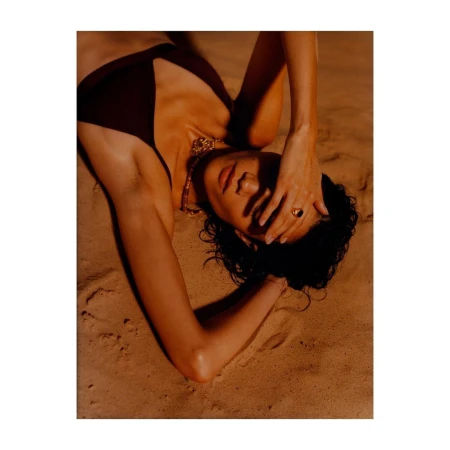Model lying on sandy terrain wearing a dark swimsuit and gold necklace with hand over forehead