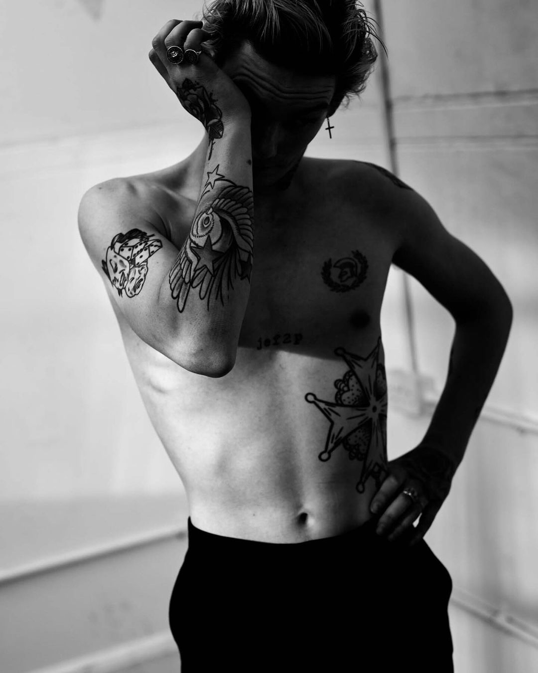 Black and white portrait of shirtless man with tattoos covering body, arm, and hand against a plain wall in an indoor setting.