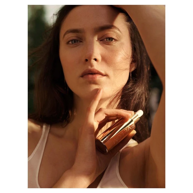 Close-up of a brunette woman holding a gold skincare tube, bathed in soft light with a blurred green outdoor background.