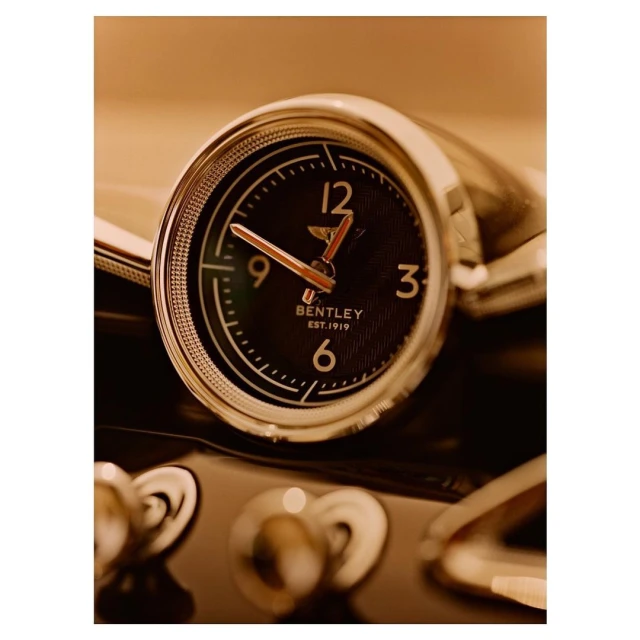 Close-up shot of a Bentley car dashboard clock; the round clock features a black face with gold-tone hands and chrome trim.