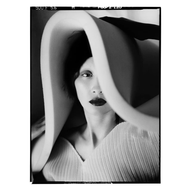 Portrait of a woman with dark lipstick wearing a cream-colored, pleated shirt and a large, sculptural hat in a black and white photo.