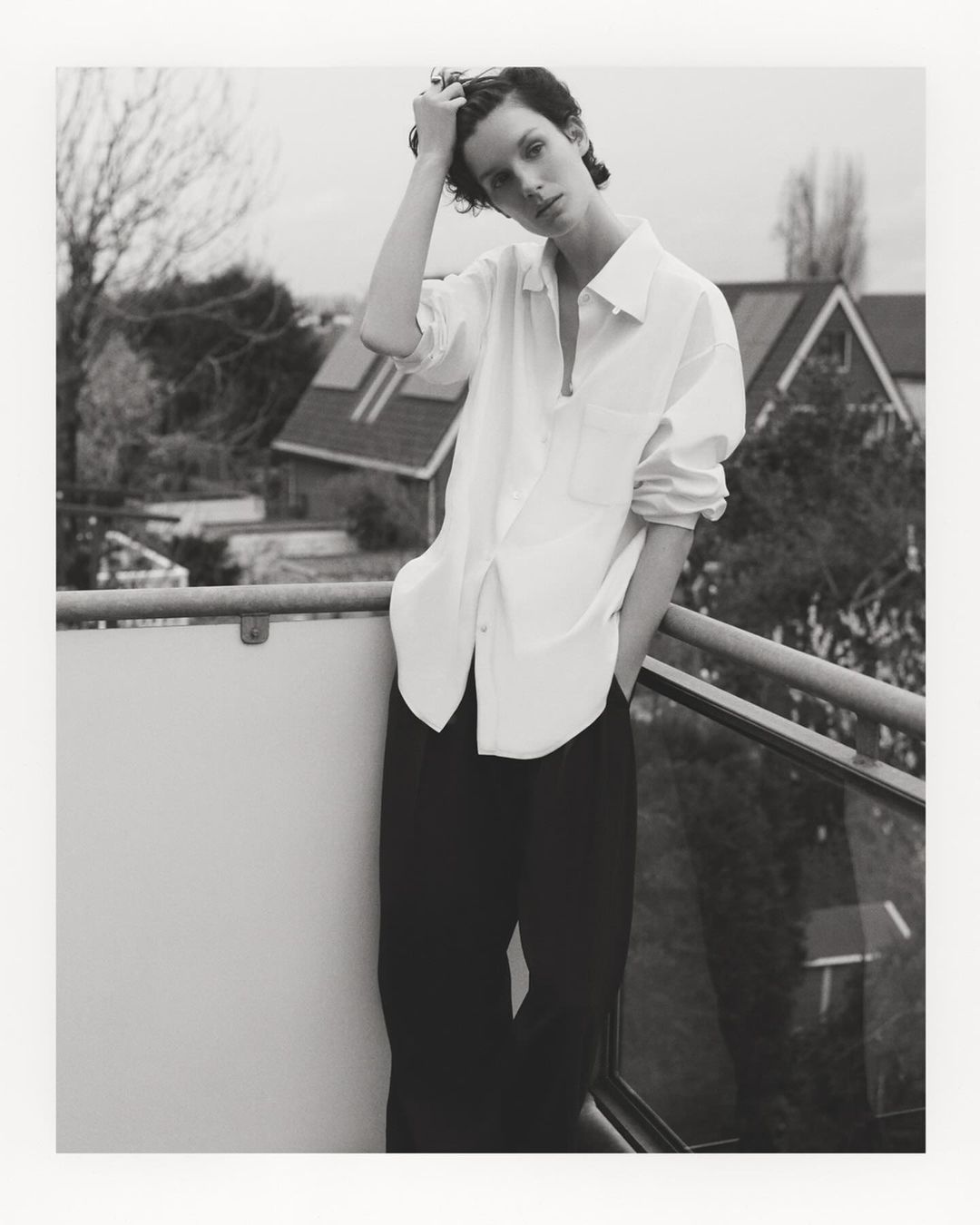 Monochrome portrait of a young woman standing on a balcony with a residential scene in the background, wearing a white shirt and dark pants.