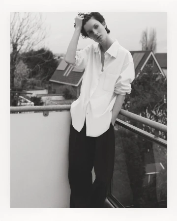 Monochrome portrait of a young woman standing on a balcony with a residential scene in the background, wearing a white shirt and dark pants.