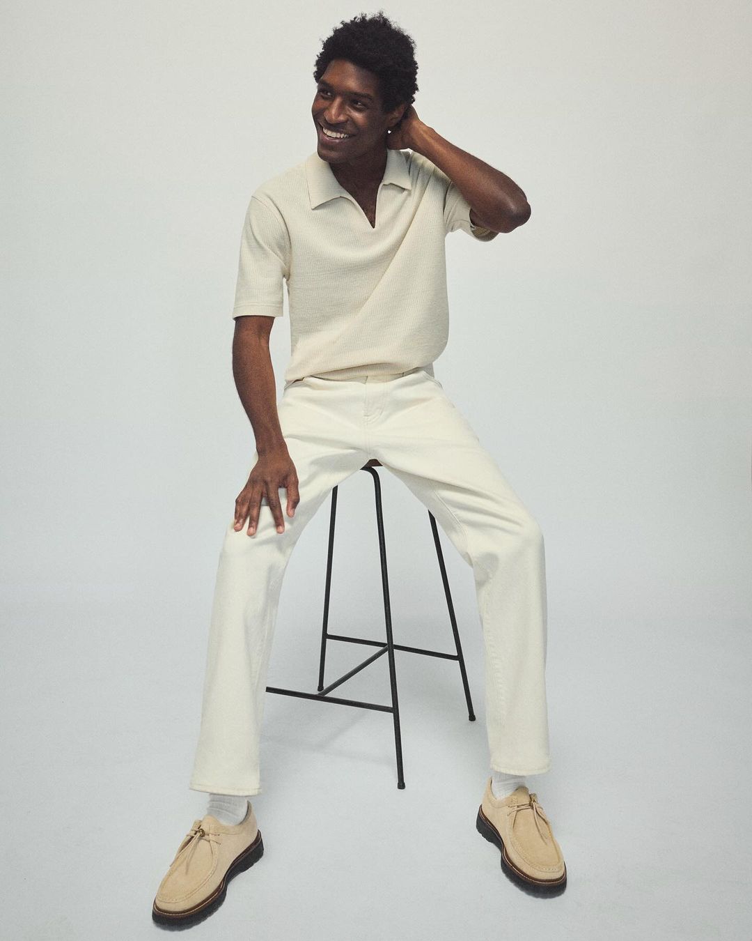 Smiling adult male model sits on a black stool, wearing beige knit polo shirt, cream-colored pants, and suede shoes in a bright studio.