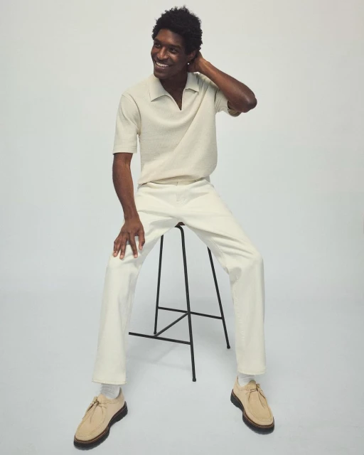 Smiling adult male model sits on a black stool, wearing beige knit polo shirt, cream-colored pants, and suede shoes in a bright studio.