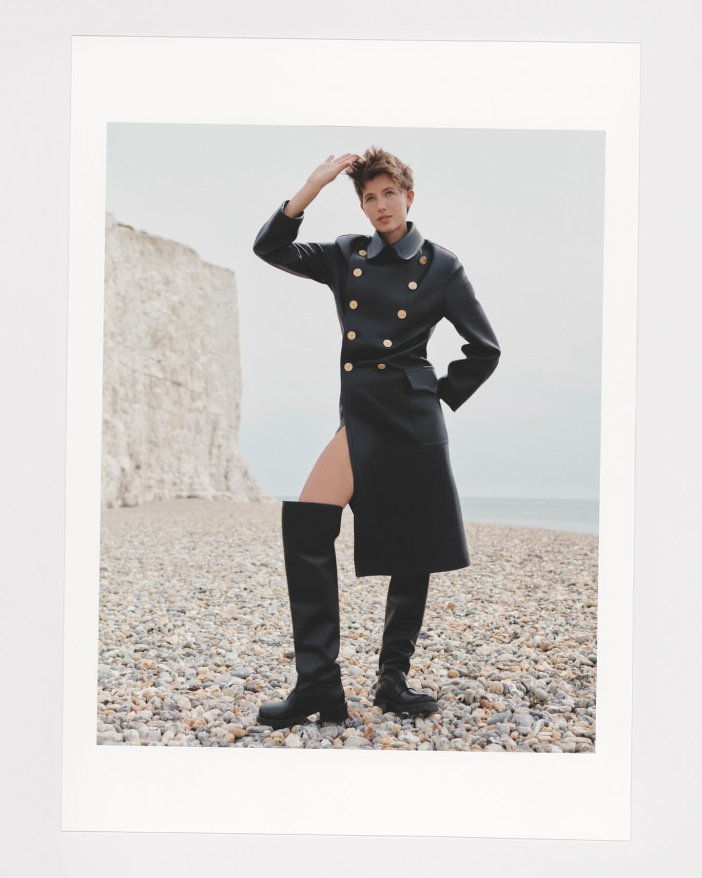 A woman in a black coat and thigh-high boots poses on a pebble beach with a chalk cliff in the background. The photo is well-lit and outdoors.