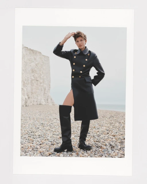 A woman in a black coat and thigh-high boots poses on a pebble beach with a chalk cliff in the background. The photo is well-lit and outdoors.