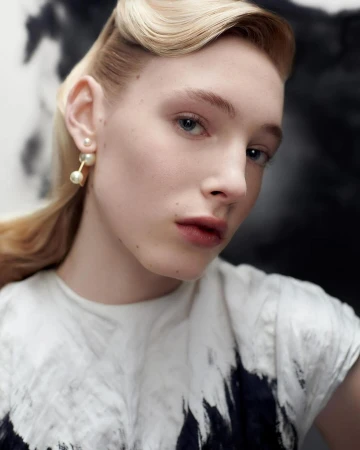 Close-up portrait of a woman wearing double pearl earrings and a patterned white top against an abstract backdrop