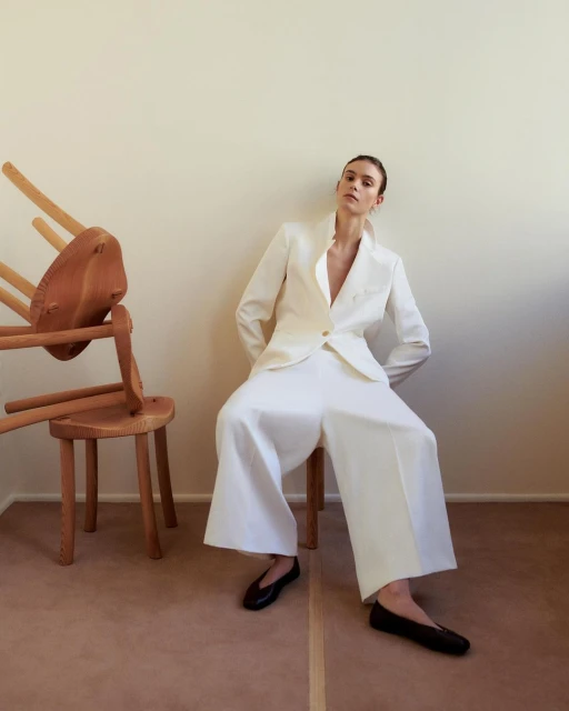 Elegant adult woman in a white suit seated by an arrangement of wooden chairs against a beige wall with a neutral floor.