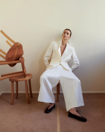 Elegant adult woman in a white suit seated by an arrangement of wooden chairs against a beige wall with a neutral floor.