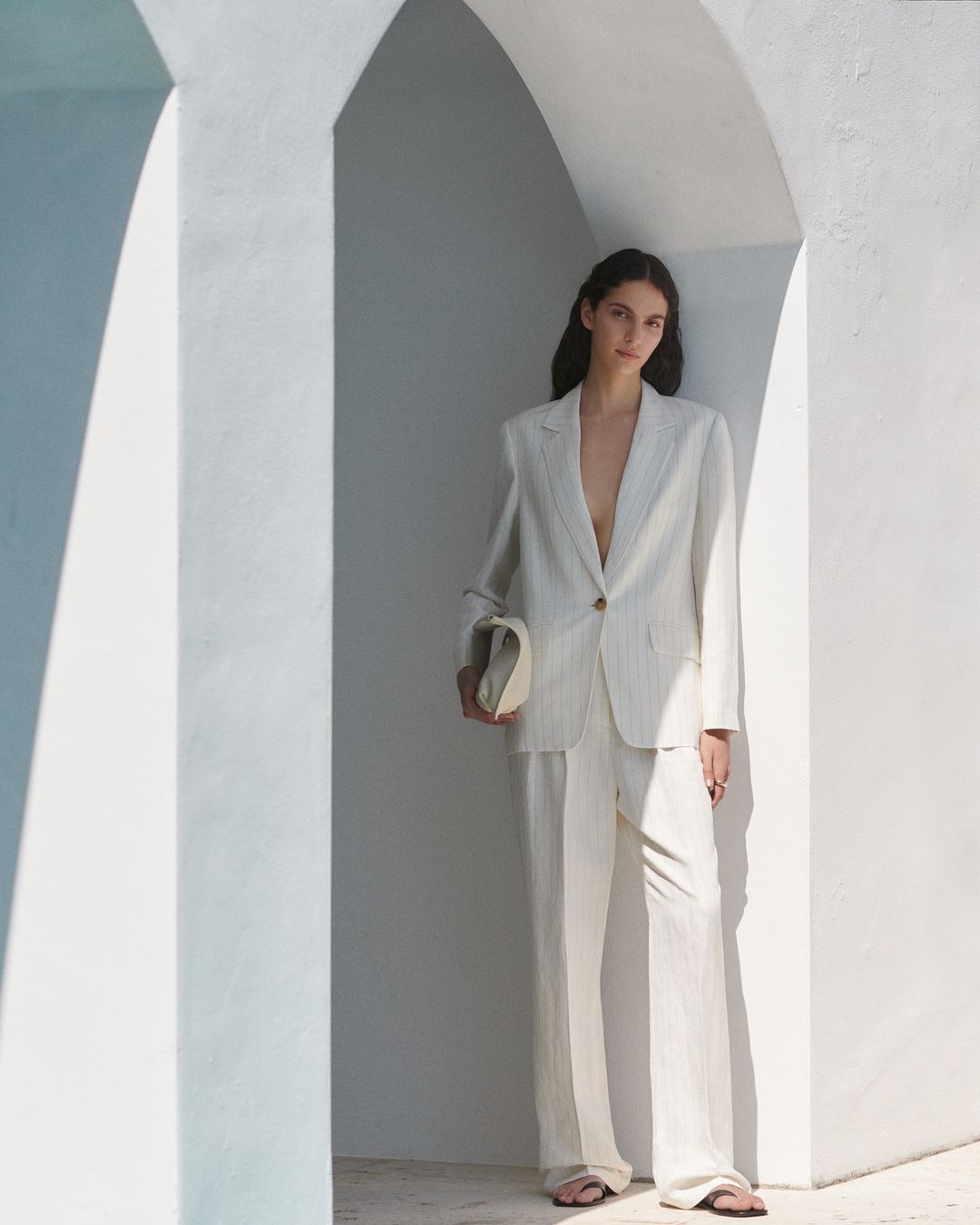 A woman in a light striped suit poses in an arched doorway, holding a clutch, with soft, diffused lighting and a minimalist aesthetic.