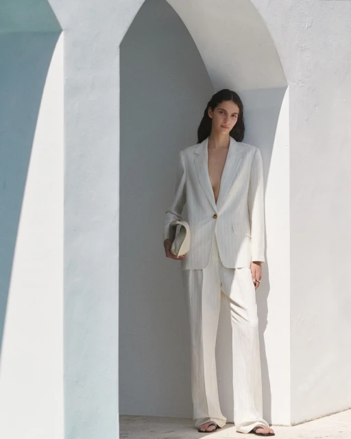 A woman in a light striped suit poses in an arched doorway, holding a clutch, with soft, diffused lighting and a minimalist aesthetic.