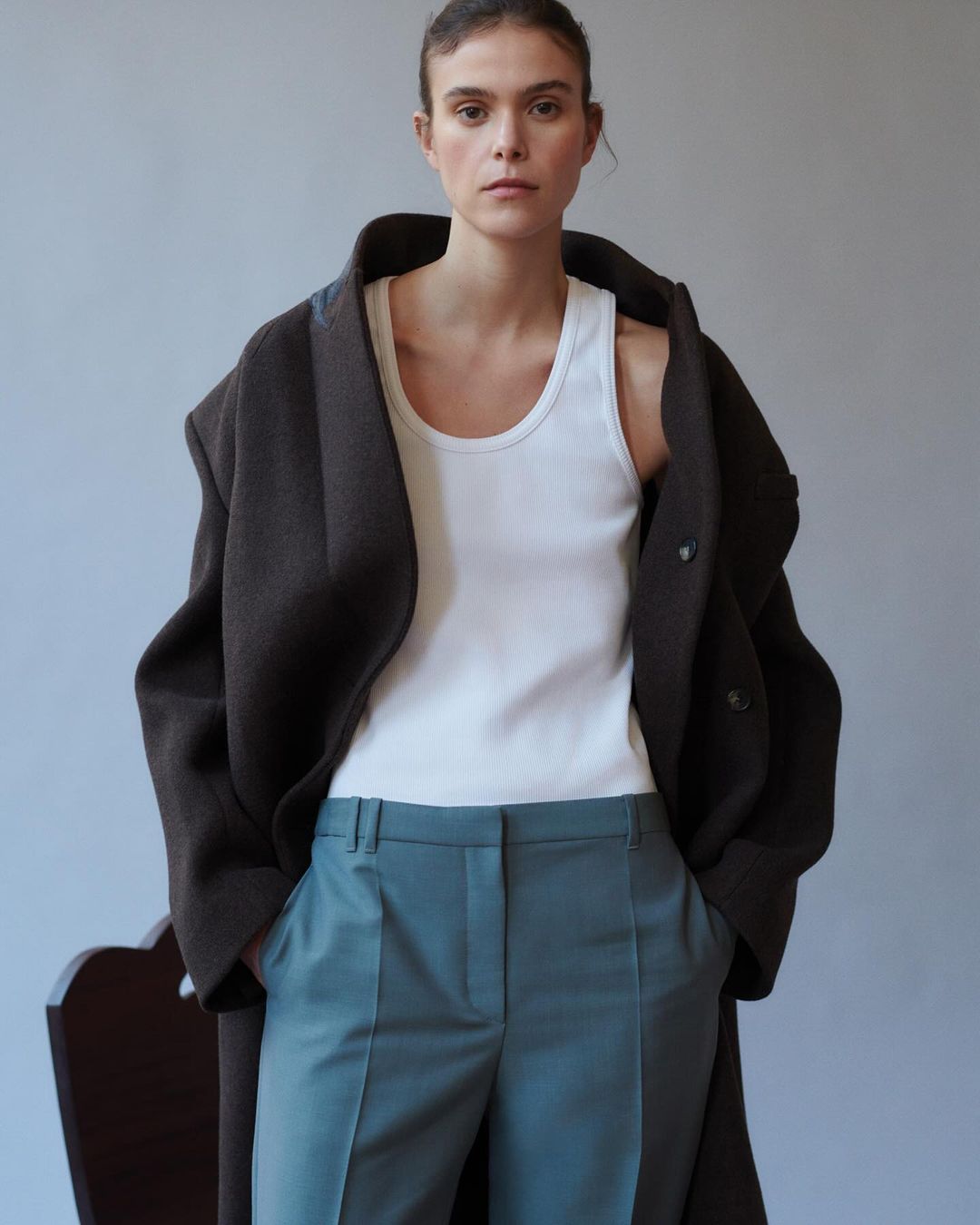 Studio shot of a woman in a white tank top, teal pants, and a dark coat. She has neutral facial expression and is indoors with natural light.