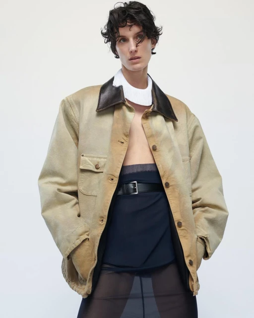 Fashion shot of a woman with short curly hair wearing a tan jacket with a dark brown collar and a black belt against a white background.