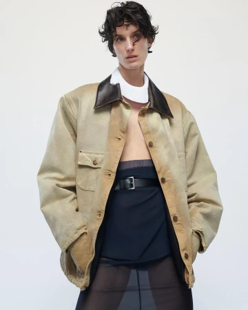 Fashion shot of a woman with short curly hair wearing a tan jacket with a dark brown collar and a black belt against a white background.