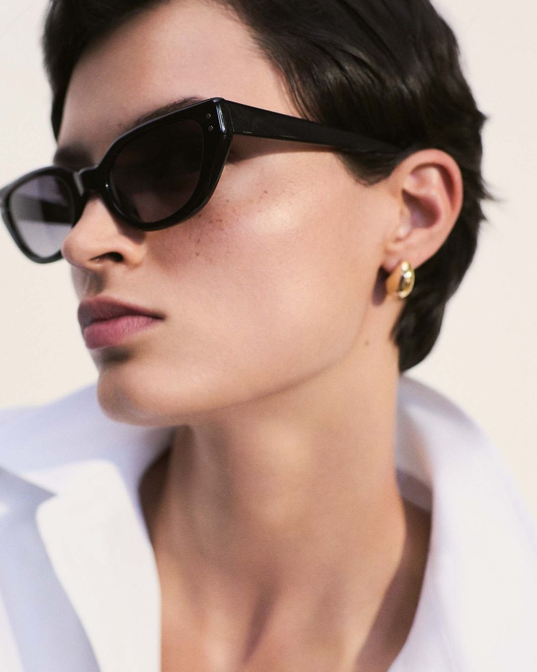 Close-up portrait of a woman wearing cat-eye sunglasses and gold hoop earrings against a neutral background, showcasing a clean aesthetic.