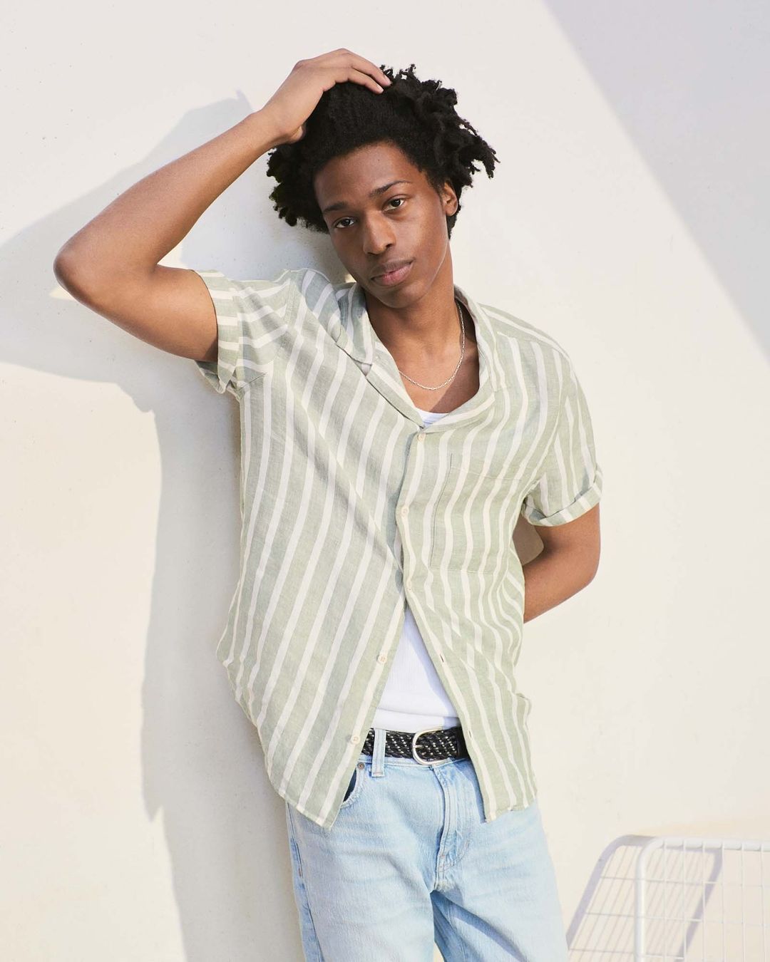 Portrait of a young adult male model with an olive striped shirt and light blue jeans, posing in a studio with a light background.