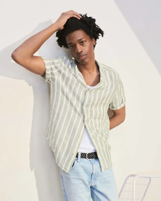 Portrait of a young adult male model with an olive striped shirt and light blue jeans, posing in a studio with a light background.