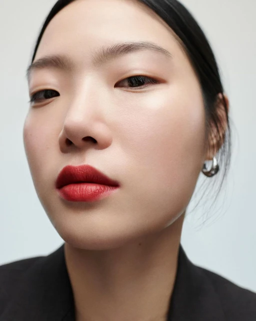 Close-up portrait of an Asian adult female model with red lipstick wearing a black jacket, captured in bright, neutral studio lighting.