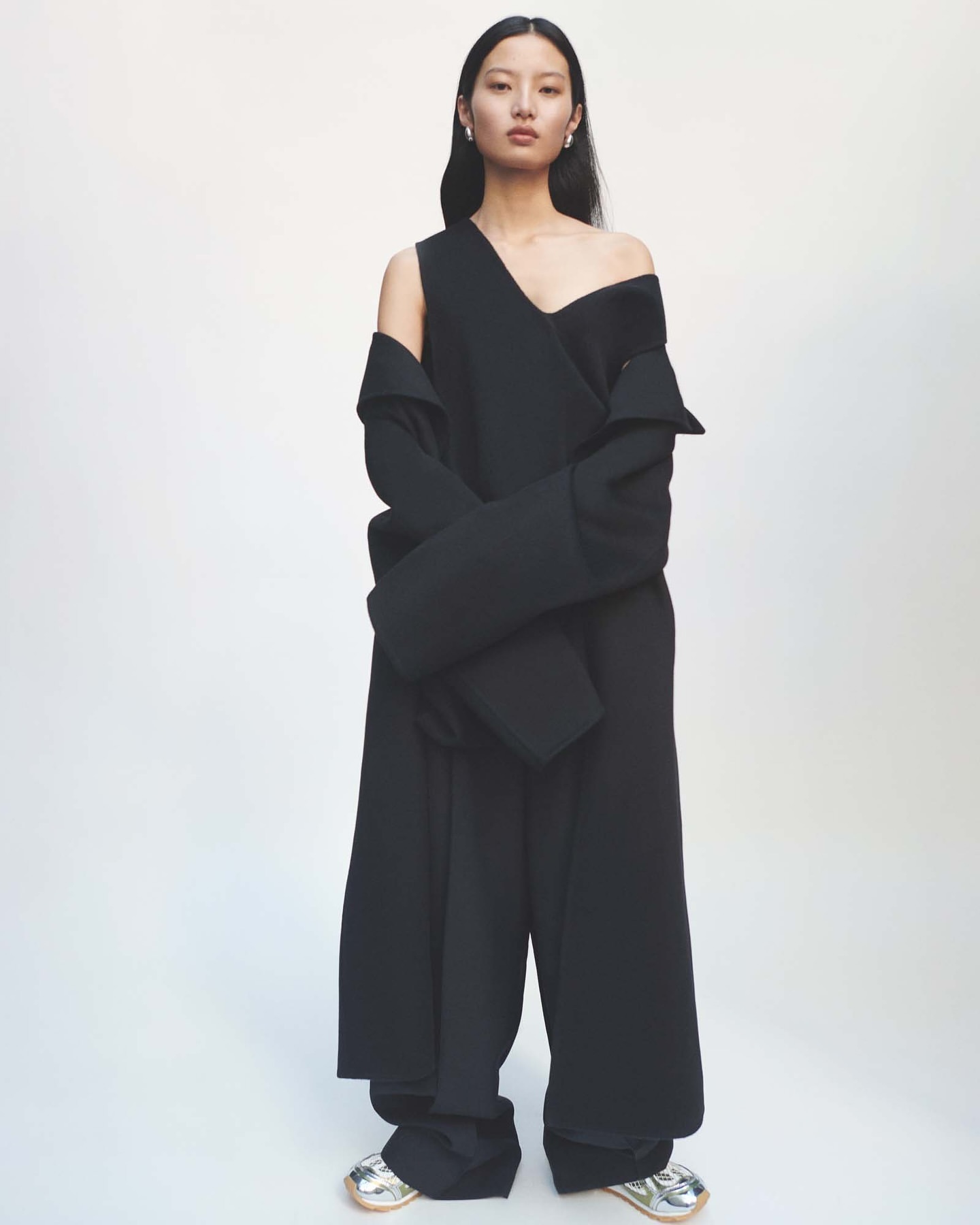 An Asian female fashion model poses in a full-length shot wearing a black, asymmetrical dress with silver shoes on a white backdrop.