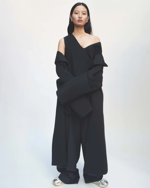 An Asian female fashion model poses in a full-length shot wearing a black, asymmetrical dress with silver shoes on a white backdrop.