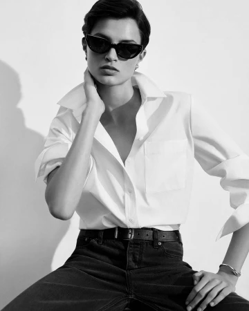 Monochrome portrait of a stylish woman in sunglasses and a white button-down shirt, exuding a confident pose against a bright background.