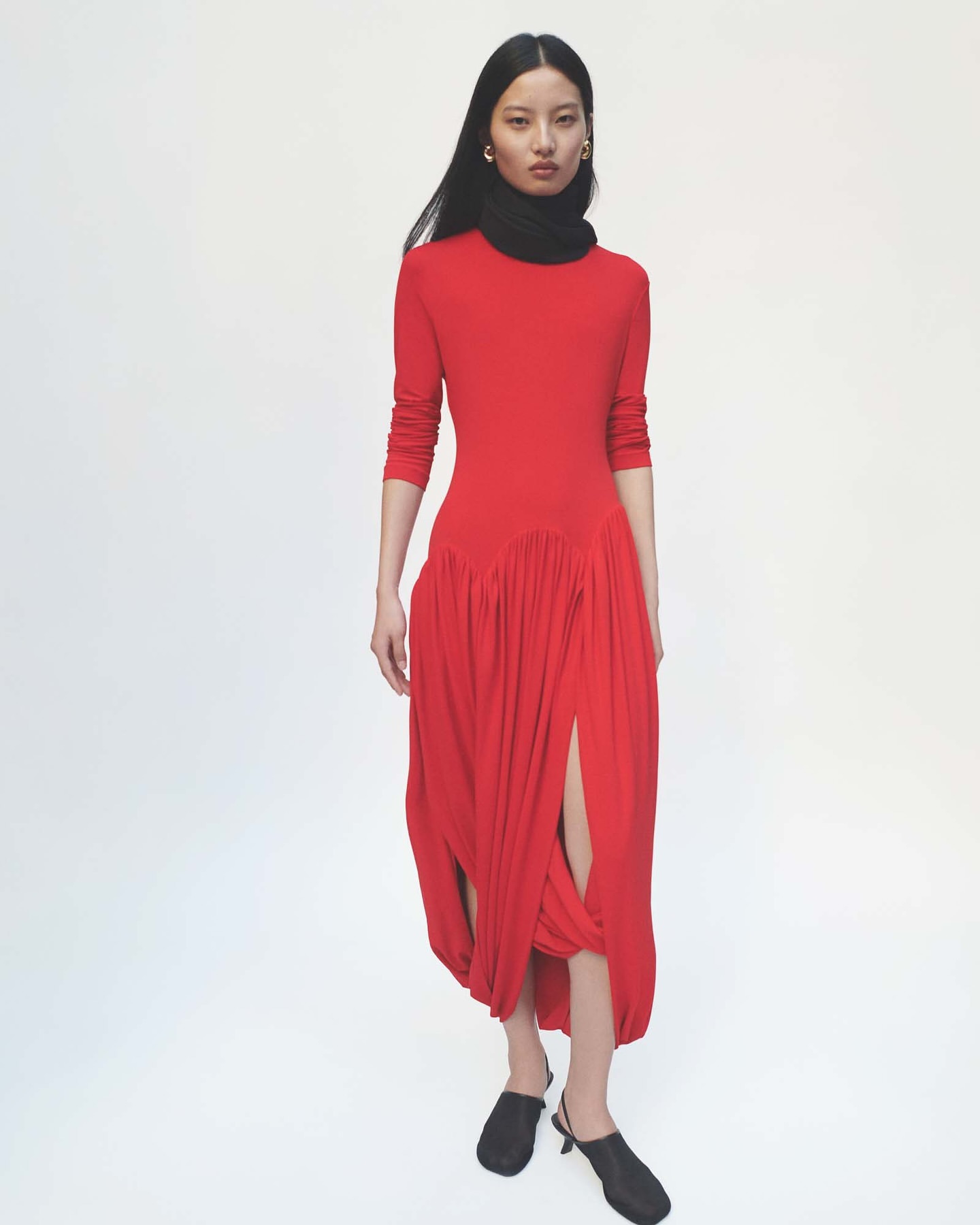 A woman poses in a full shot in a vibrant red dress with a black scarf, standing against a stark white backdrop, showcasing fashion elegance.