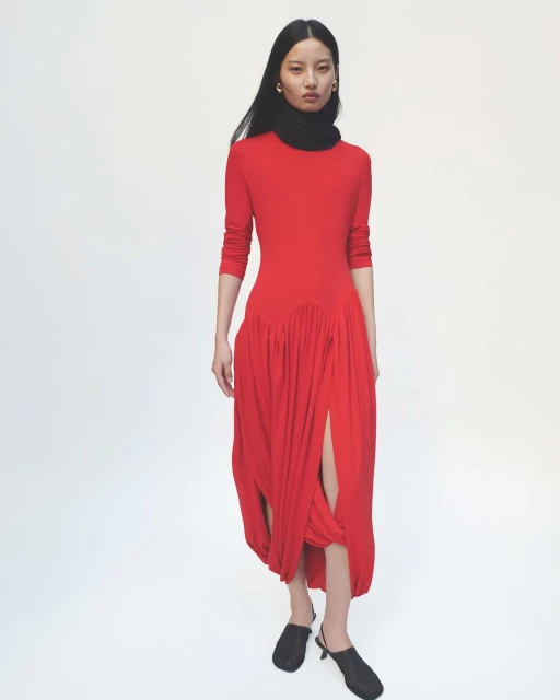 A woman poses in a full shot in a vibrant red dress with a black scarf, standing against a stark white backdrop, showcasing fashion elegance.