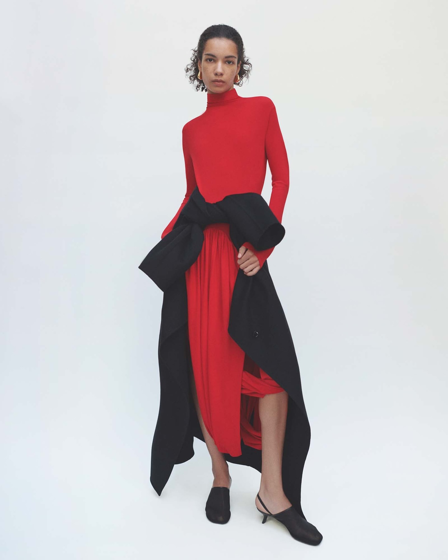 Model wearing a red turtleneck top with a black coat tied around the waist against white background
