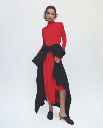 Model wearing a red turtleneck top with a black coat tied around the waist against white background